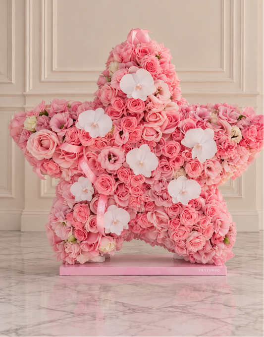 A large star-shaped floral arrangement made of pink roses and white orchids on a pink base, displayed indoors against a cream paneled wall and marble floor.