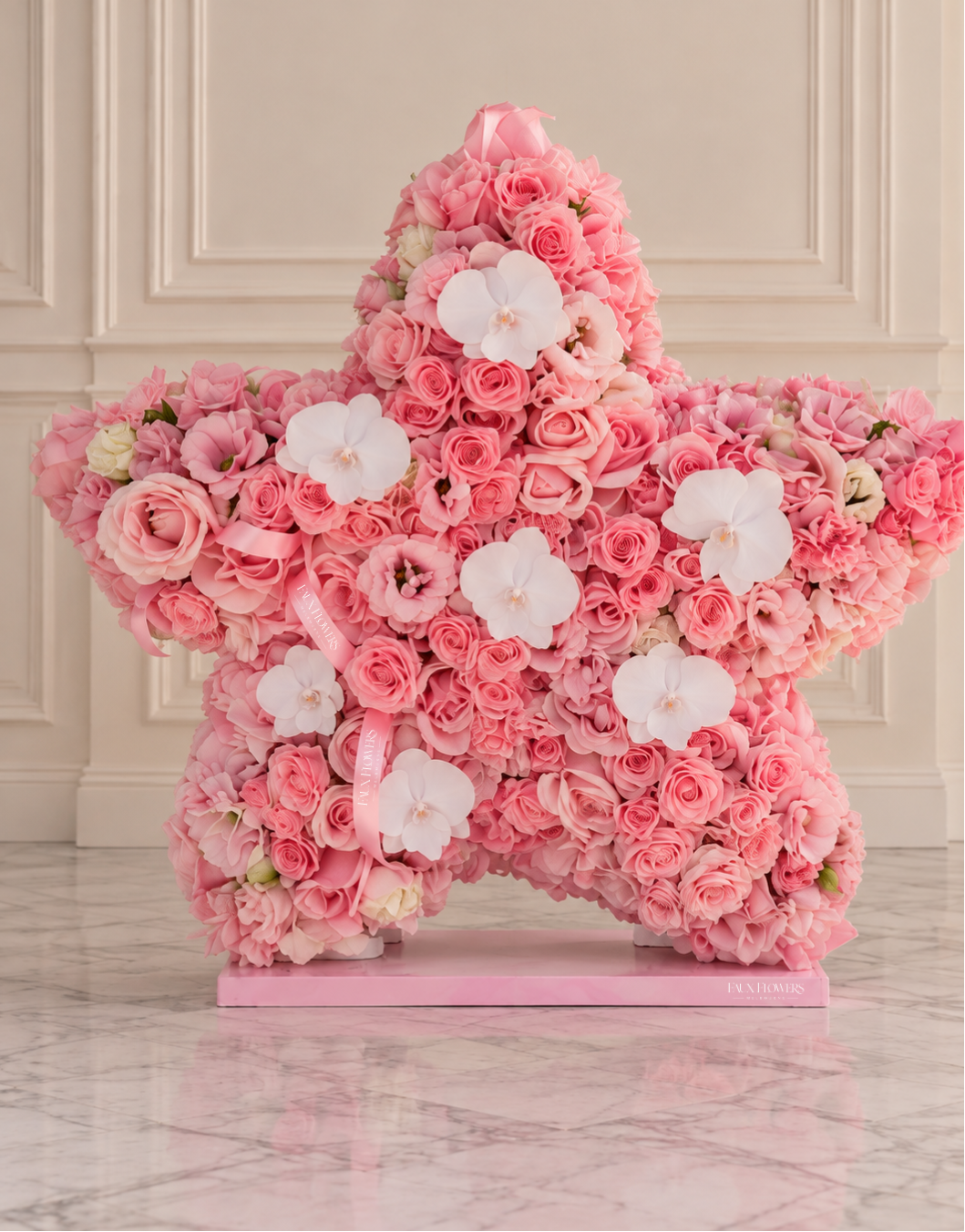 A large star-shaped floral arrangement made of pink roses and white orchids on a pink base, displayed indoors against a cream paneled wall and marble floor.